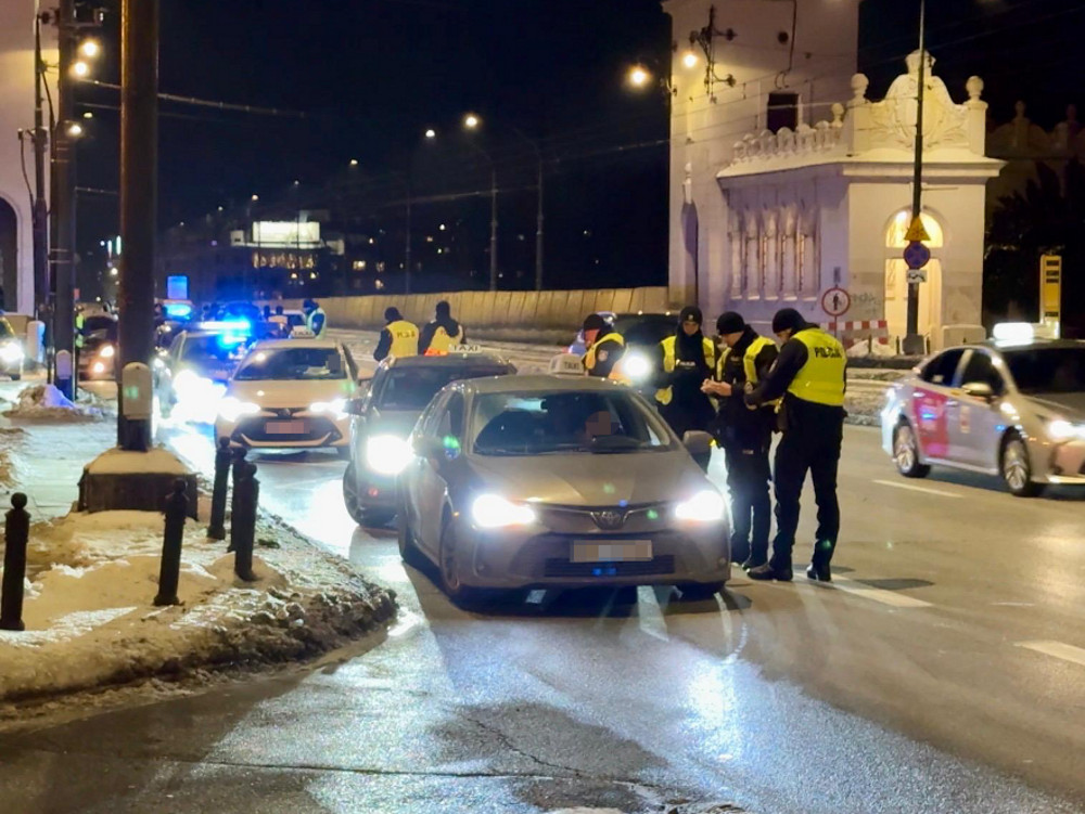Grupa policjantów w odblaskowych kamizelkach kontroluje kierowcę zatrzymanego samochodu na oświetlonej ulicy nocą.