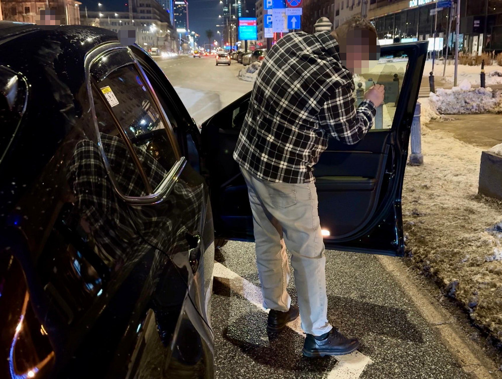 Mężczyzna w koszuli w kratę sprawdza dokumenty w otwartym samochodzie na oświetlonej ulicy w nocy.