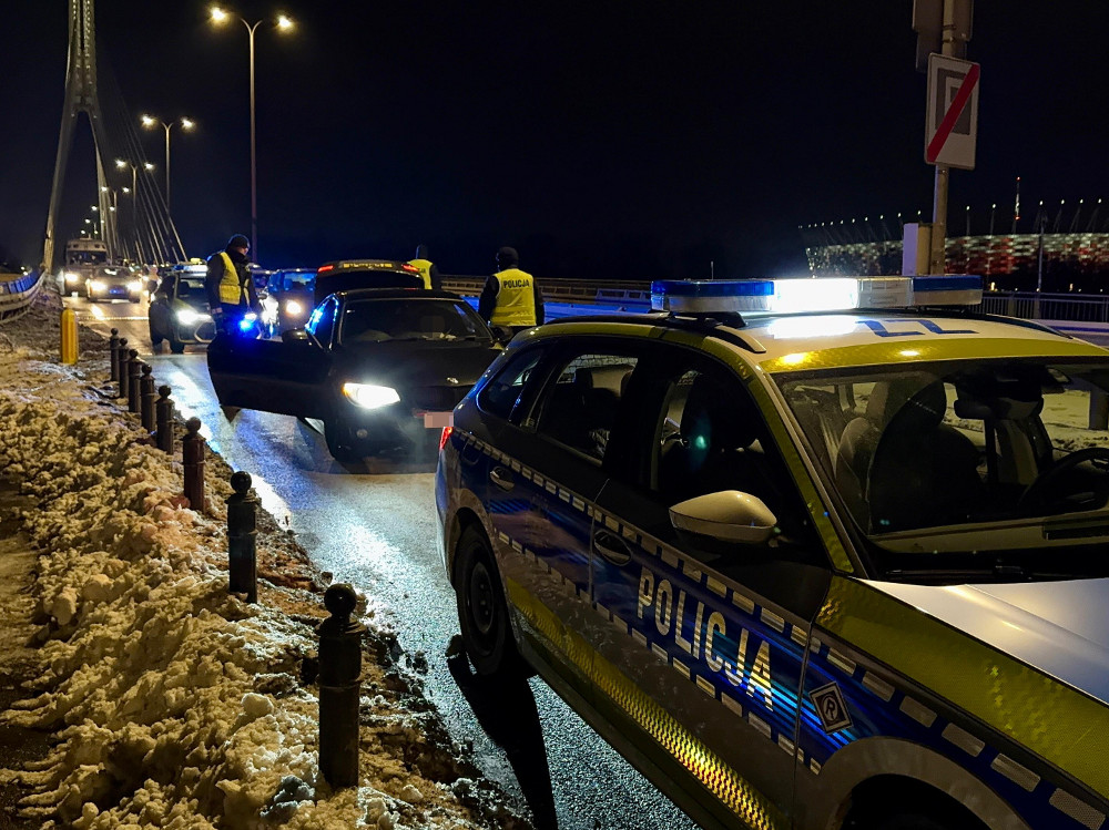 Policjanci w odblaskowych kamizelkach kontrolują pojazdy na oświetlonej drodze w nocy, obok leżącego śniegu.