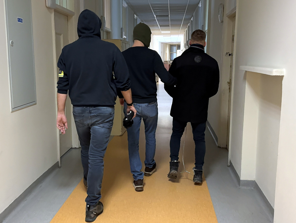 Trzech mężczyzn idzie korytarzem w budynku, jeden z nich ma rękę na ramieniu drugiego, a trzeci trzyma kamerę.