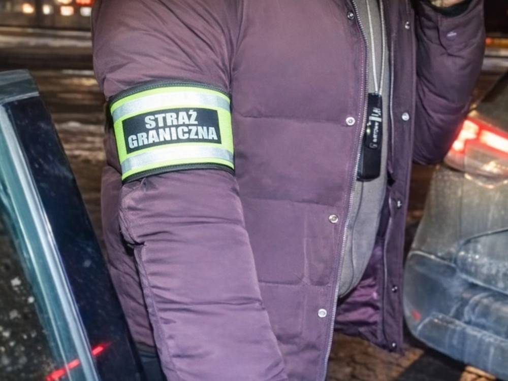 Mężczyzna w kurtce z odblaskową opaską "STRAŻ GRANICZNA" rozmawia przez telefon obok samochodu.