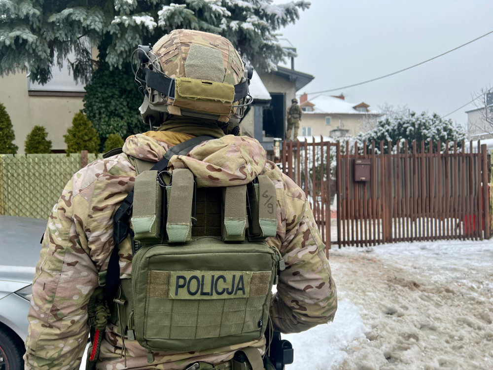 Mężczyzna w kamuflażu, z plecakiem oznaczonym napisem "POLICJA", stoi na zaśnieżonej ulicy przed ogrodzeniem.