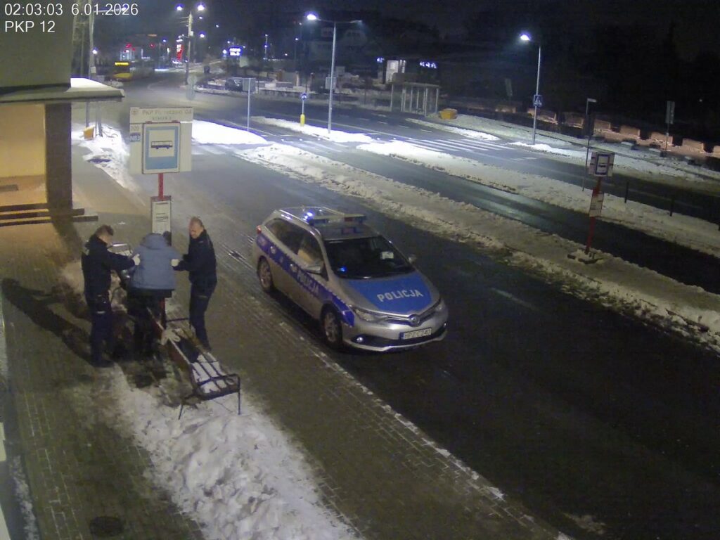 Dwaj policjanci pomagają mężczyźnie w niebieskiej kurtce, siedzącym na ławce przy przystanku autobusowym w nocy.