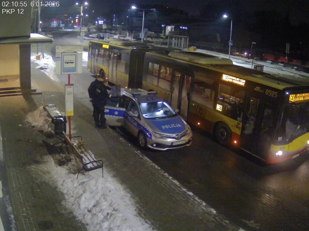 Policjant rozmawia z kierowcą radiowozu obok przystanku, w tle stoi autobus na oświetlonej ulicy.