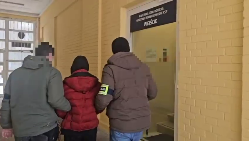 Trzy osoby w odzieży zimowej idą w kierunku drzwi z napisem "WEJŚCIE" w jasnym korytarzu.