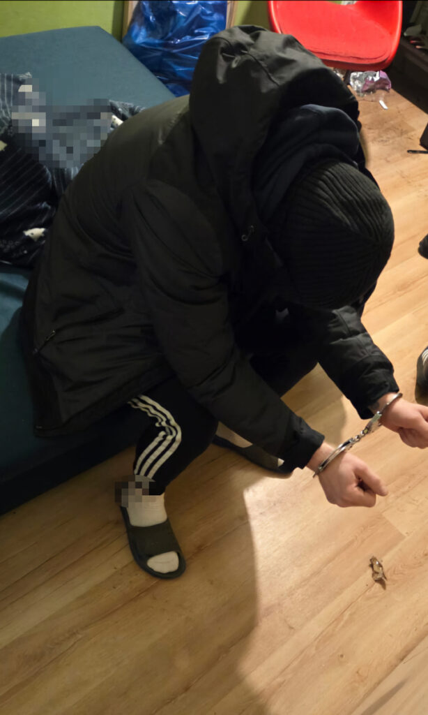 Mężczyzna w czarnej kurtce i czapce siedzi na podłodze, z rękami skuty w kajdankach, w nieformalnym wnętrzu.