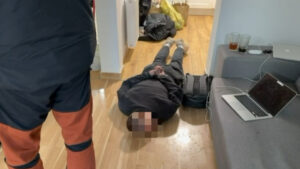 Mężczyzna leży na podłodze z rękami związanymi, podczas gdy inna osoba stoi obok w pomieszczeniu mieszkalnym.