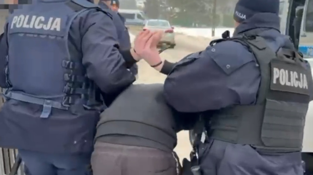 Policjanci w mundurach zatrzymują mężczyznę, który jest unieruchomiony i ma założone kajdanki.