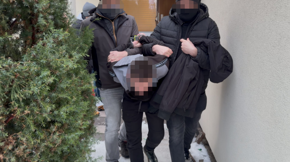 Grupa mężczyzn w ciemnych ubraniach prowadzi osobę w szarej bluzie na zewnątrz budynku, w otoczeniu zielonych krzewów.