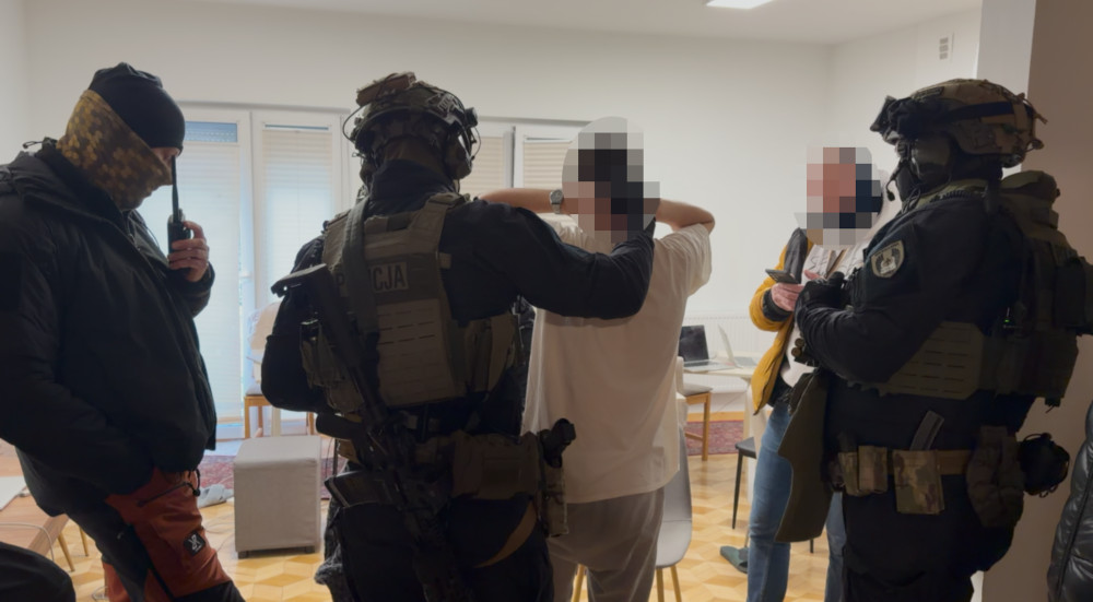 Czterech uzbrojonych funkcjonariuszy policji przeszukuje mieszkanie, kontrolując osobę w białej koszulce.