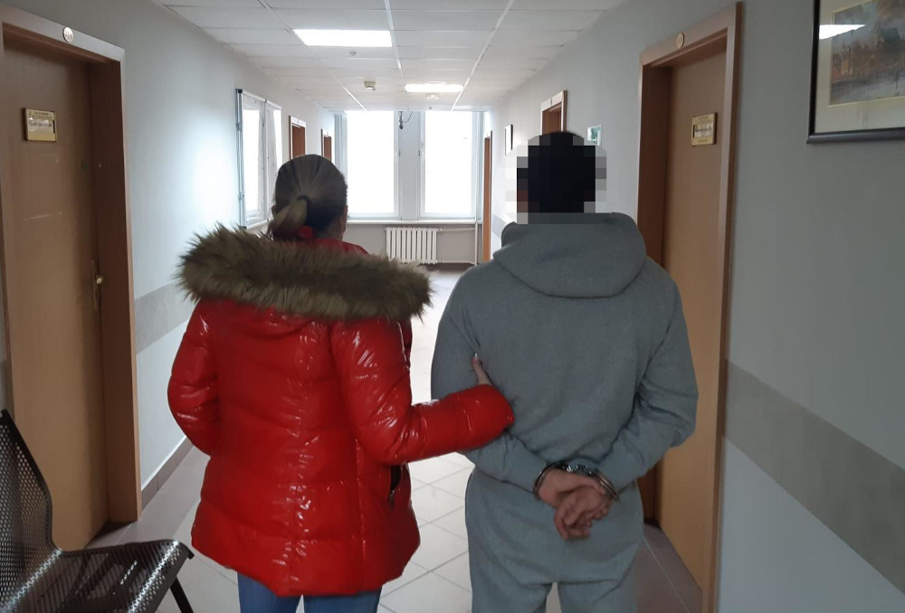 Kobieta w czerwonej kurtce prowadzi mężczyznę w szarym dresie z rękami związanymi z tyłu, w korytarzu budynku.