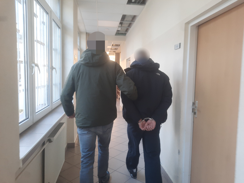 Mężczyzna w płaszczu prowadzi aresztowanego mężczyznę w kajdankach korytarzem w budynku.