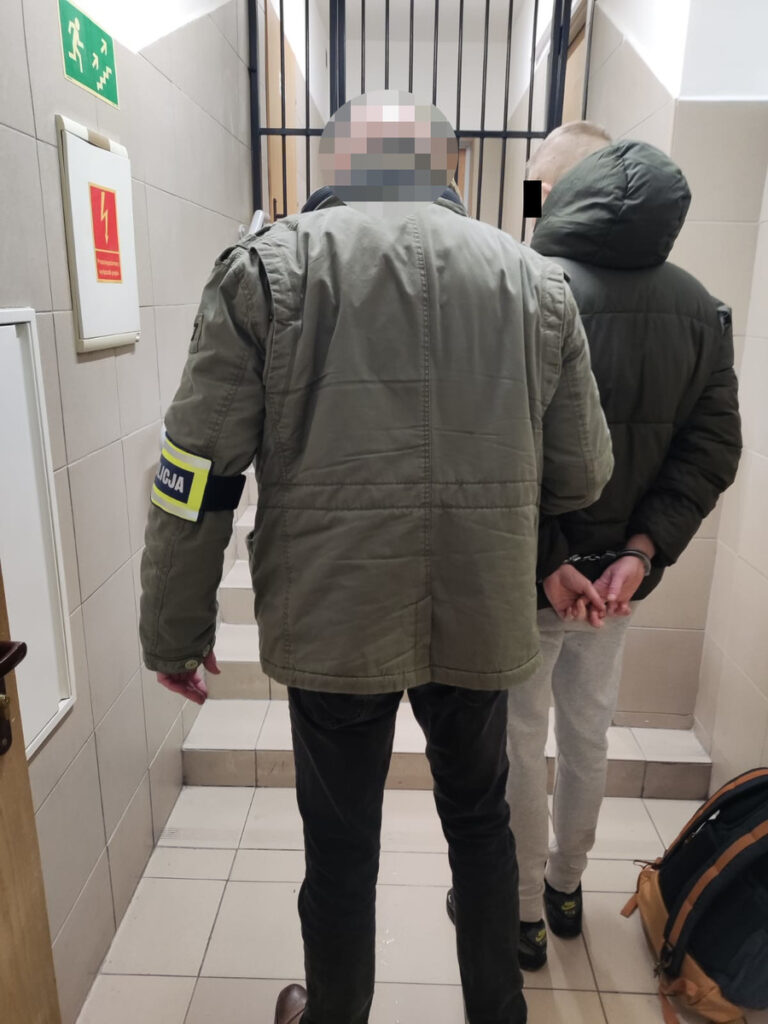 Funkcjonariusz w mundurze prowadzi zatrzymanego mężczyznę w kajdankach w korytarzu budynku.