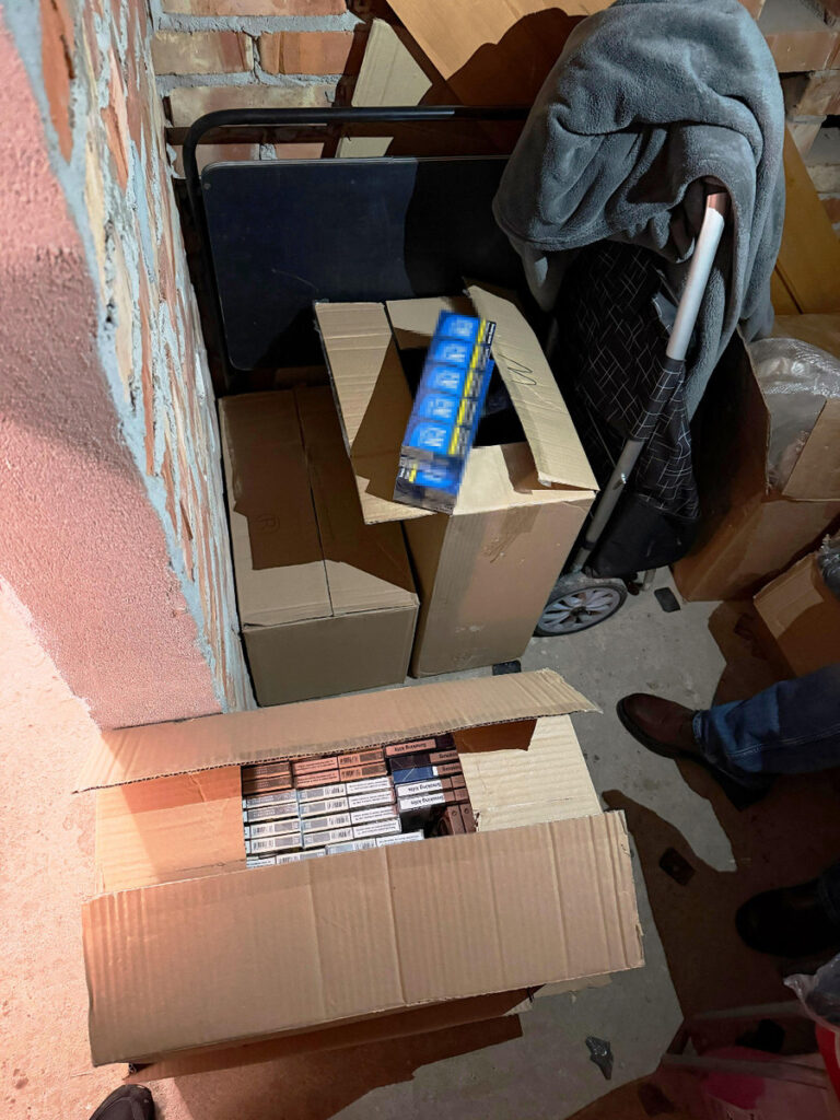 W kącie pomieszczenia stoją kartony, w jednym z nich widoczne są paczki papierosów, obok wózek z kocem.