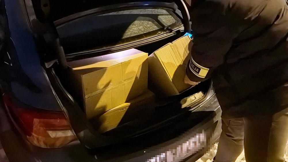 Mężczyzna w kurtce wkłada kartonowe pudła do bagażnika ciemnego samochodu w nocy.