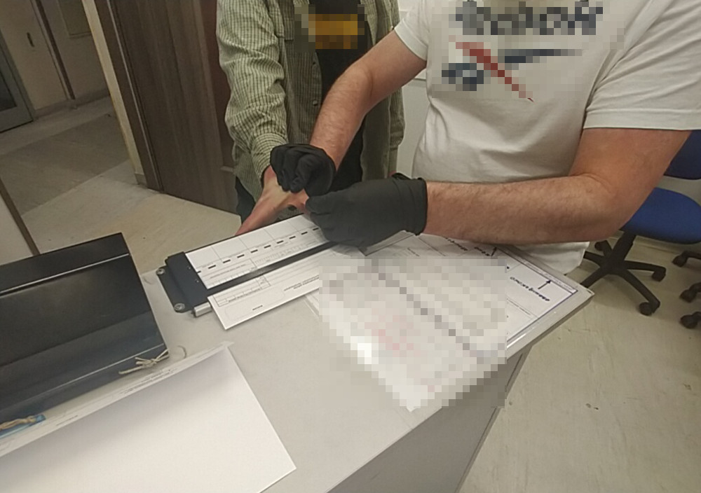 Mężczyzna w rękawiczkach dotyka urządzenia elektronicznego na biurku, podczas gdy inna osoba pomaga mu w tym procesie.