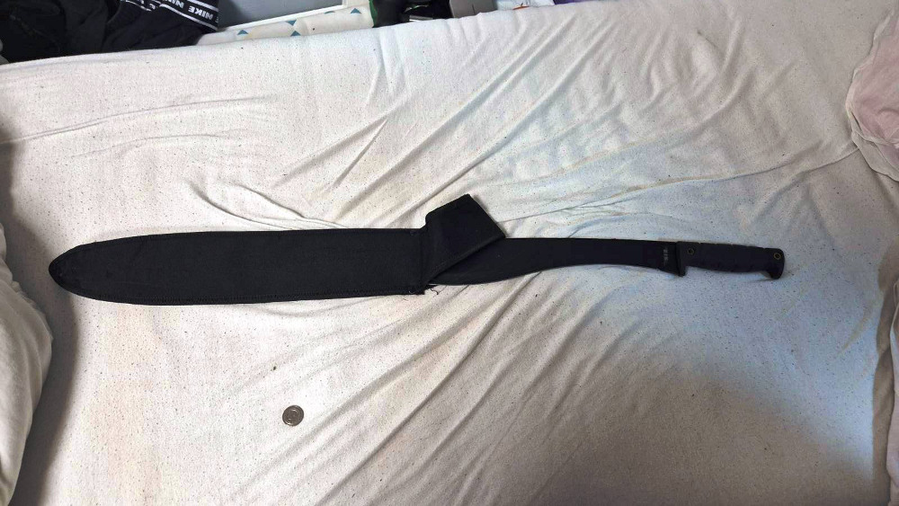 Czarny nóż w osłonie leży na białej pościeli obok monety.