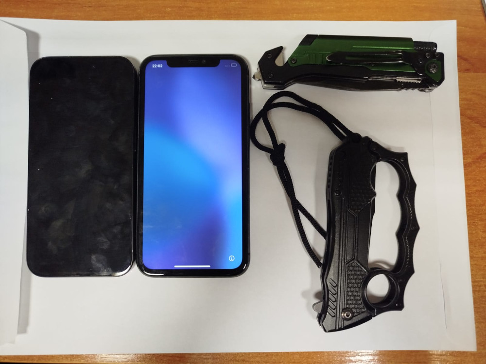 Na białym tle leżą dwa smartfony, jeden z czarną obudową, a drugi włączony z niebieskim ekranem, obok znajduje się czarny nóż z uchwytem oraz zielony multitool.
