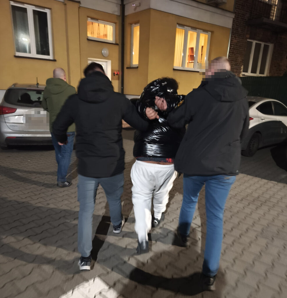 Dwaj mężczyźni w ciemnych kurtkach prowadzą trzecią osobę w połyskującej kurtce, na chodniku w pobliżu budynku mieszkalnego.