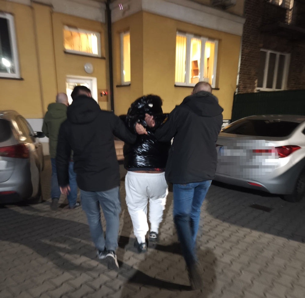 Dwaj mężczyźni w czarnych kurtkach prowadzą osobę w błyszczącej kurtce przez parking nocą.