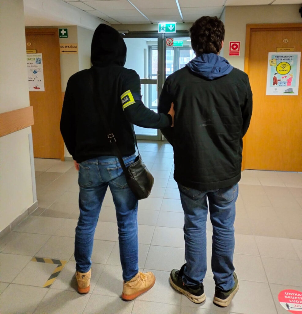 Mężczyzna w czarnej bluzie z kapturem prowadzi drugiego mężczyznę korytarzem w budynku.