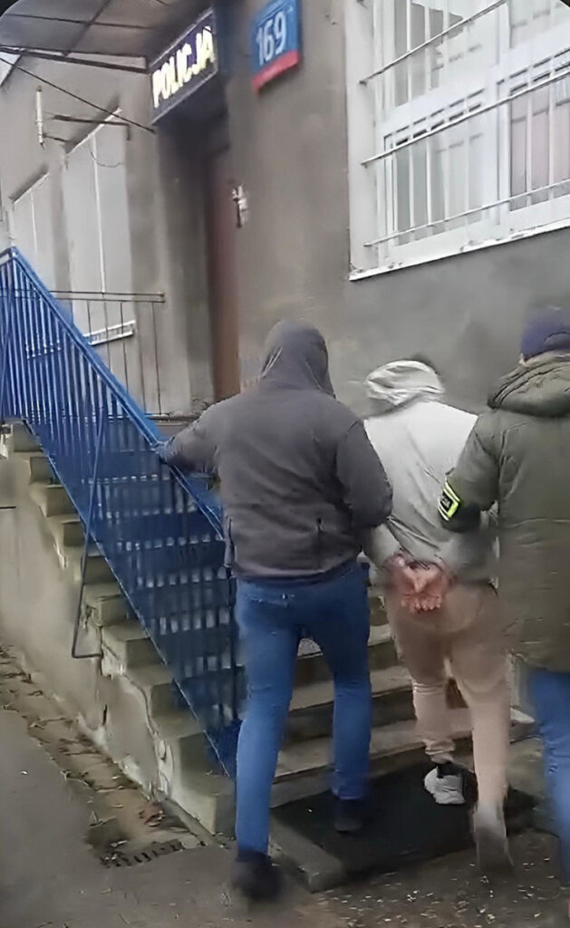 Mężczyzna w szarym dresie prowadzony przez policjanta w mundurze w kierunku wejścia do budynku policji.