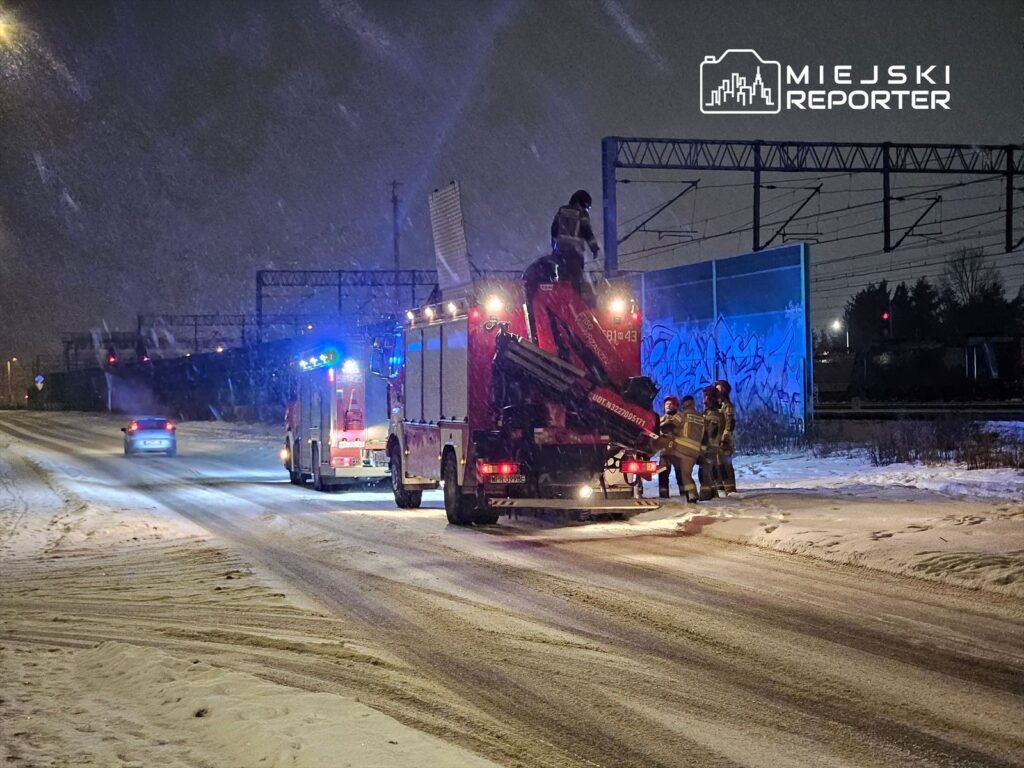 Strażacy pracują przy wozie strażackim na zaśnieżonej drodze w nocy, w tle widoczne pociągi i graffiti na murze.