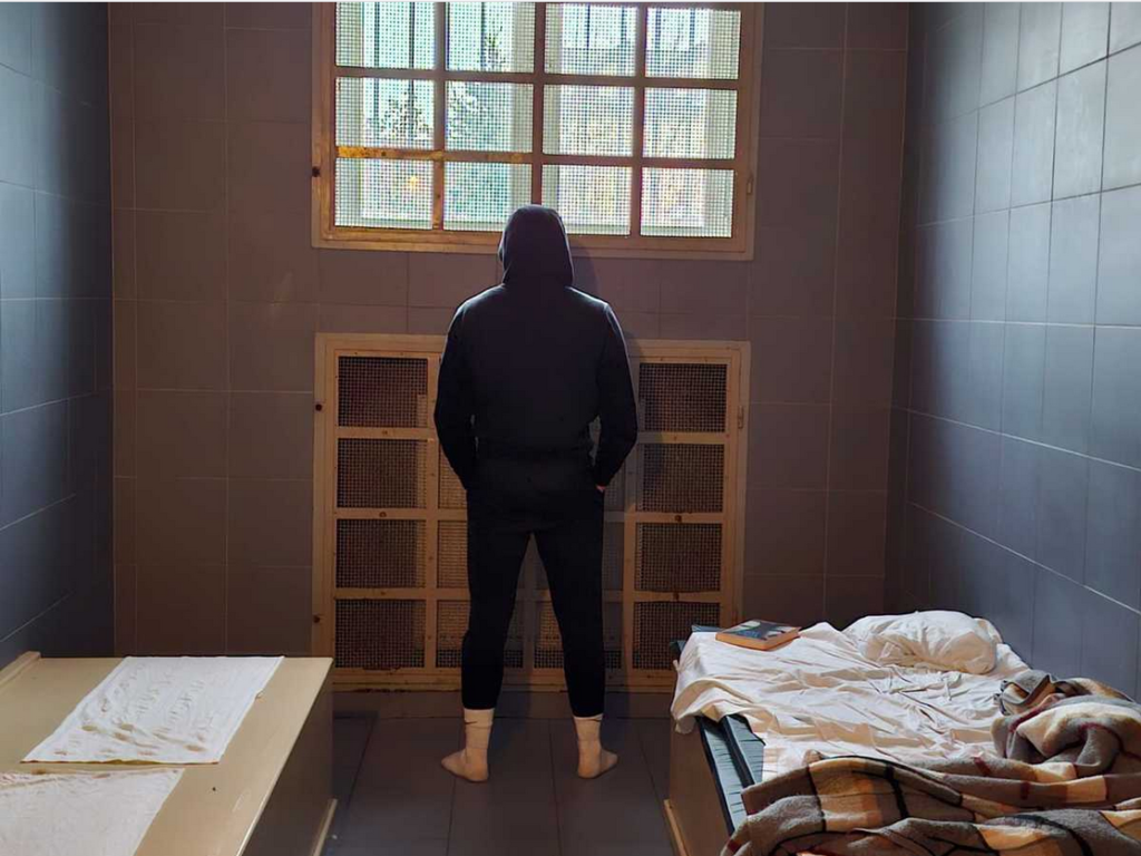 Mężczyzna w czarnej bluzie stoi plecami do okna w małym pomieszczeniu z łóżkami i pościelą.