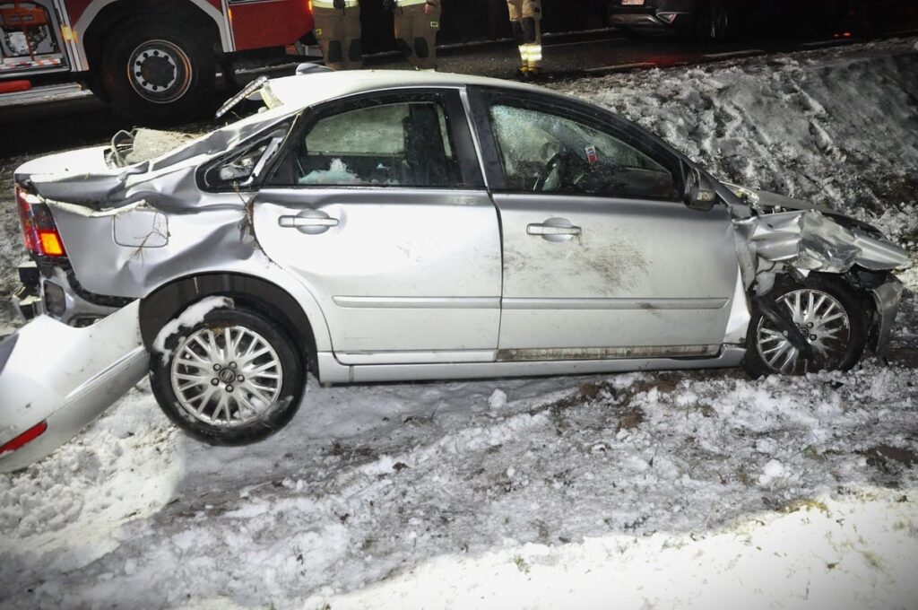 Uszkodzony samochód osobowy leży w śniegu obok drogi, a w tle widać samochody ratunkowe i strażaków.
