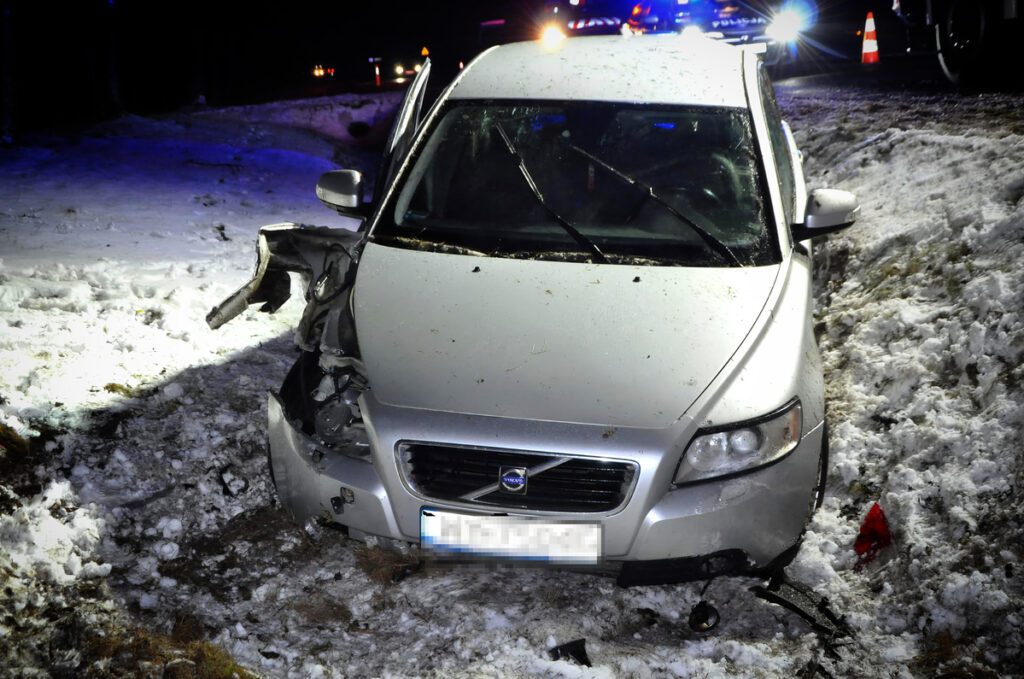 Srebrny samochód osobowy z uszkodzoną przednią częścią stoi w śniegu obok drogi, w tle widoczne światła policyjne.