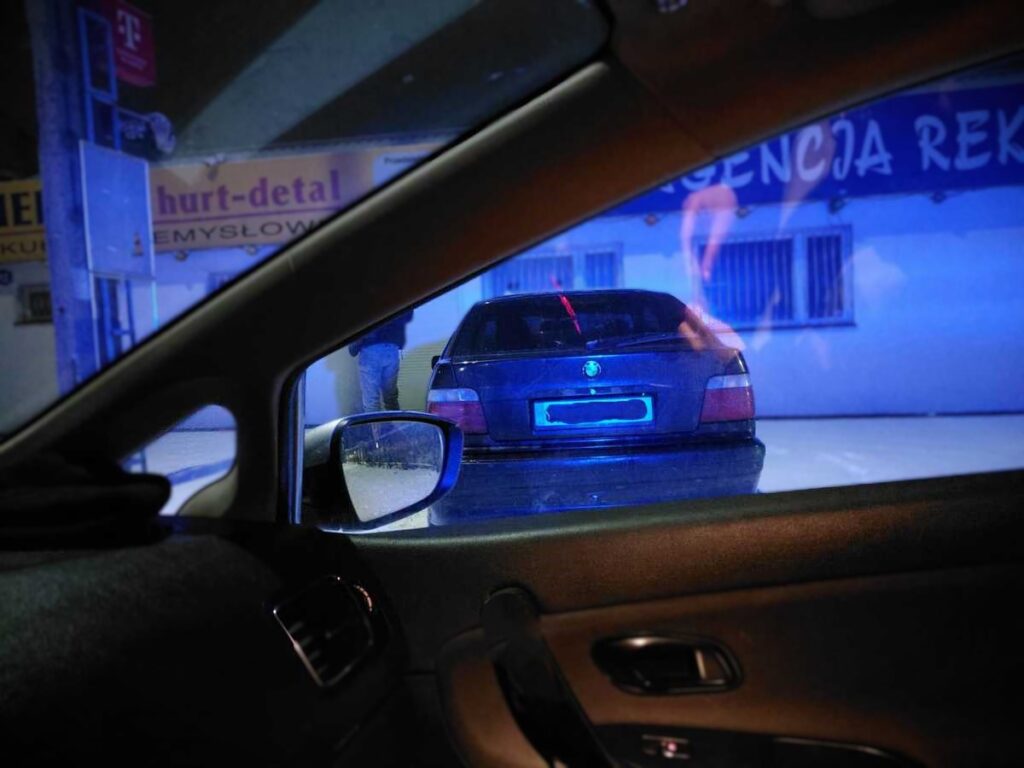 Mężczyzna w niebieskim świetle policyjnym stoi obok czarnego samochodu zaparkowanego przed budynkiem.