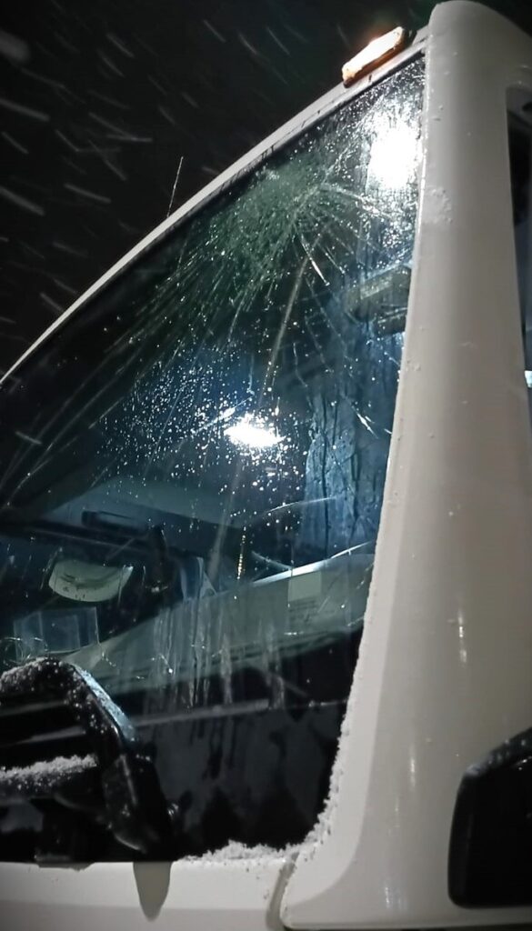 Pęknięta szyba kabiny pojazdu ciężarowego pokryta śniegiem w nocy, z odbiciem światła latarni.