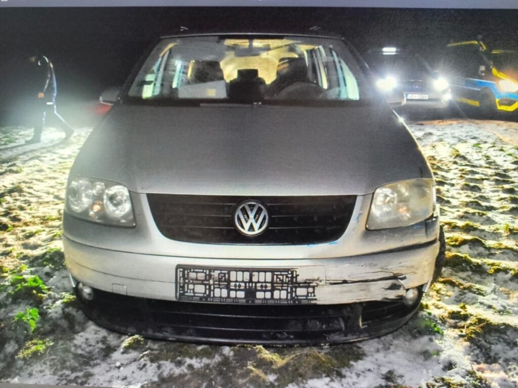 Jasnoszary Volkswagen stoi na oświetlonym piasku, z widocznymi uszkodzeniami z przodu; w tle widać mężczyznę idącego w stronę pojazdu.