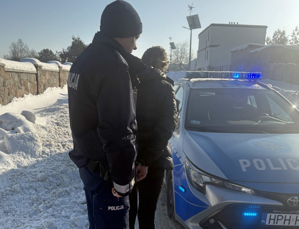 Mężczyzna w czarnej kurtce zatrzymywany przez policjanta obok radiowozu na zaśnieżonej ulicy.