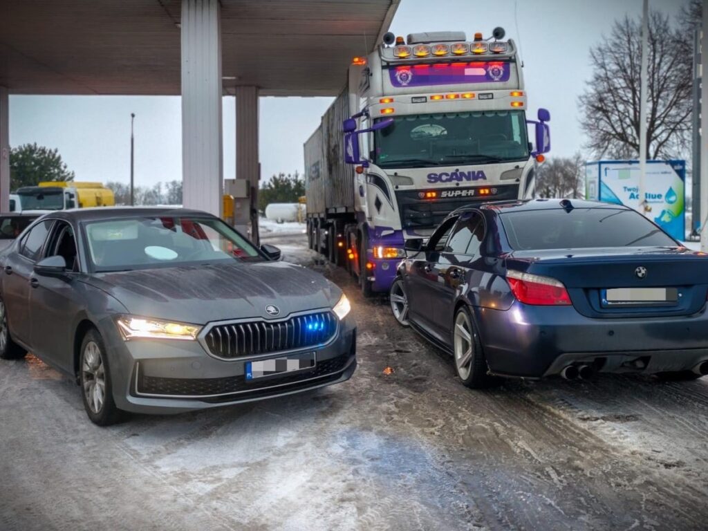Skrzyżowanie dróg z policyjnym samochodem osobowym z niebieskim światłem oraz ciężarówką na stacji paliw.