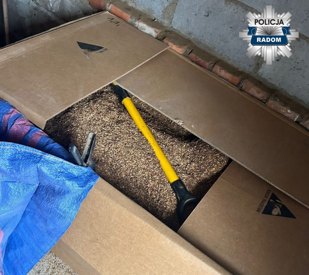 Skrzynia z otwartą pokrywą, wypełniona drobnym materiałem, obok łopata z żółtą rączką na niebieskiej plandece.