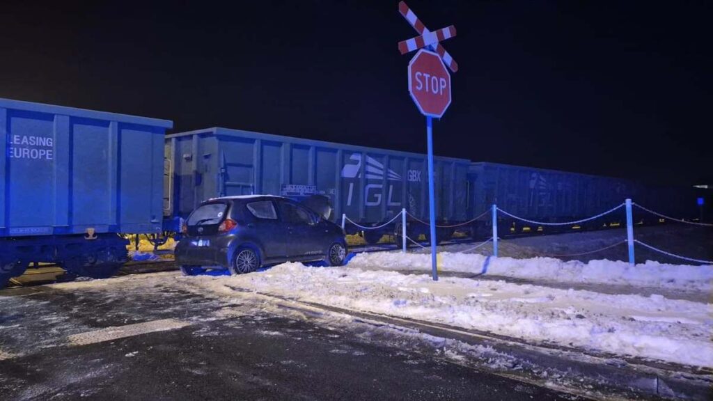 Samochód osobowy stoi na torach kolejowych, obok zespołu niebieskich wagonów towarowych, w otoczeniu śniegu i znaku stop.