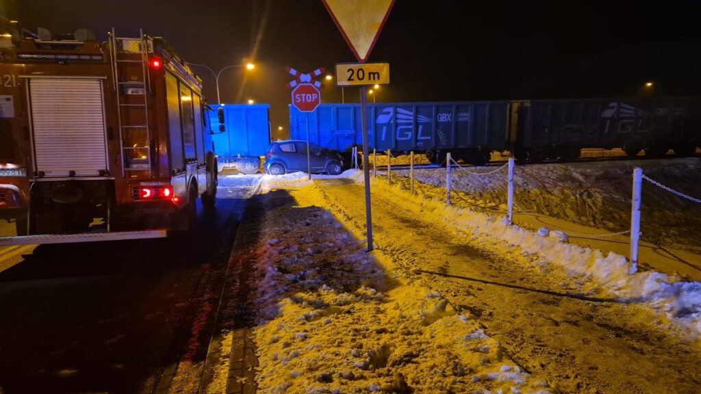 W nocy widać wypadek drogowy, gdzie samochód osobowy zderzył się z pociągiem towarowym na zaśnieżonej drodze. Obok stoi wóz strażacki.