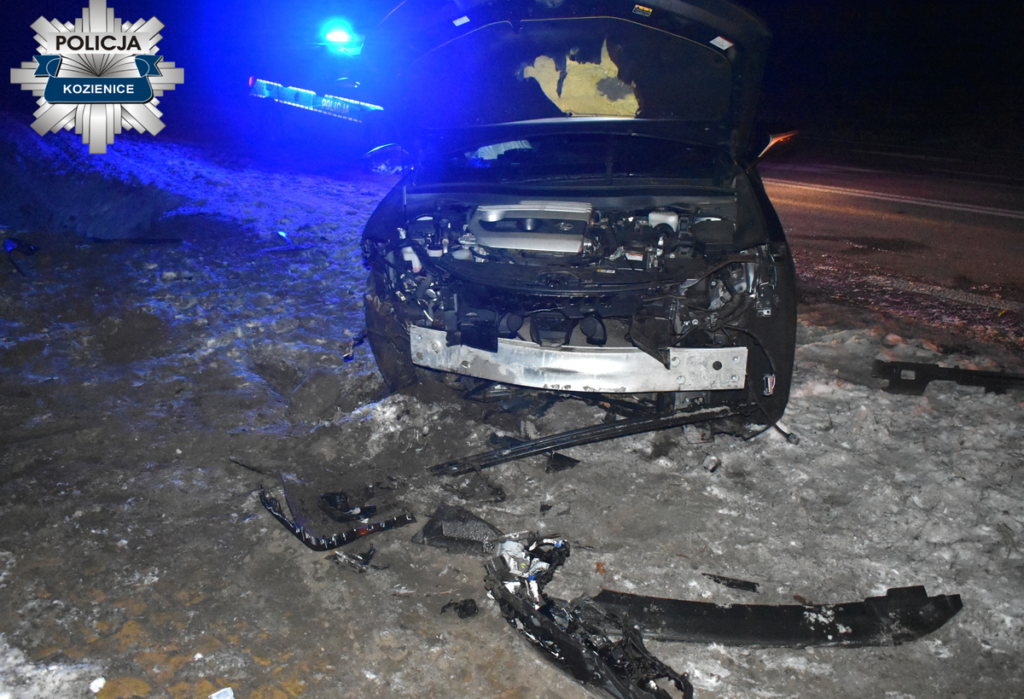 Uszkodzony samochód osobowy z otwartą maską leży na poboczu drogi, w tle widać policyjny radiowóz z włączonymi sygnałami świetlnymi.