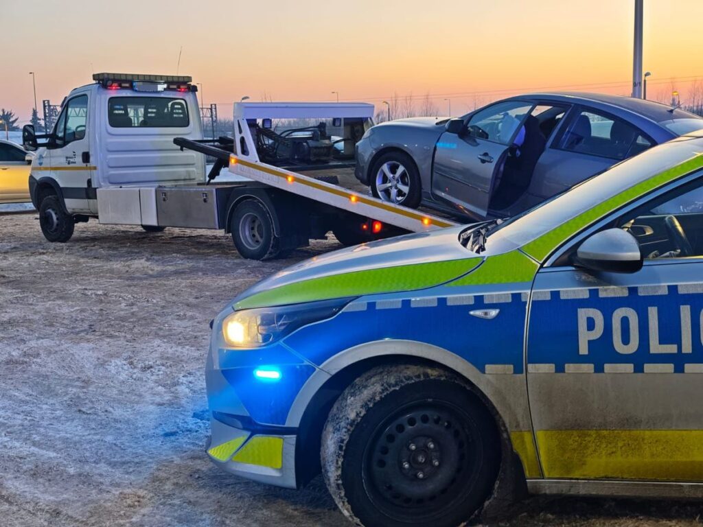 Policyjny samochód stoi na zaśnieżonym parkingu, podczas gdy laweta podnosi pojazd osobowy.