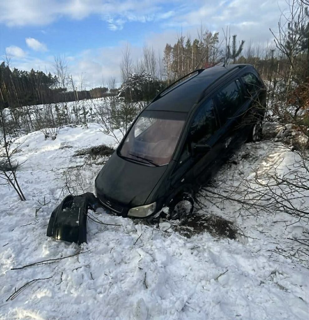 Czarny minivan utknął w śniegu, z przodu odłamana część zderzaka, otoczony drzewami w leśnym terenie.