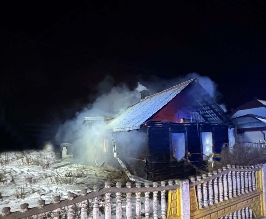 Dom z dymem i ogniem na dachu, otoczony przez zarośla i ogrodzenie, w nocy.