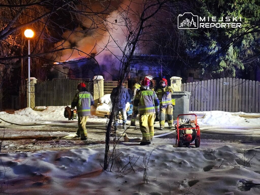 Grupa strażaków w mundurach omawia działania ratunkowe w pobliżu płonącego budynku w zimowej scenerii.