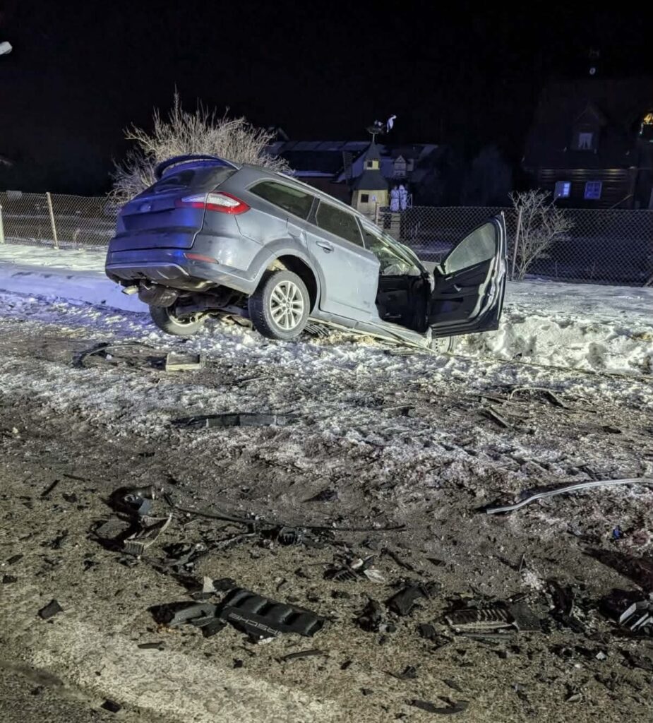Samochód osobowy leży na boku na poboczu drogi, otwarta drzwi, wokół rozrzucone fragmenty pojazdu i śnieg.