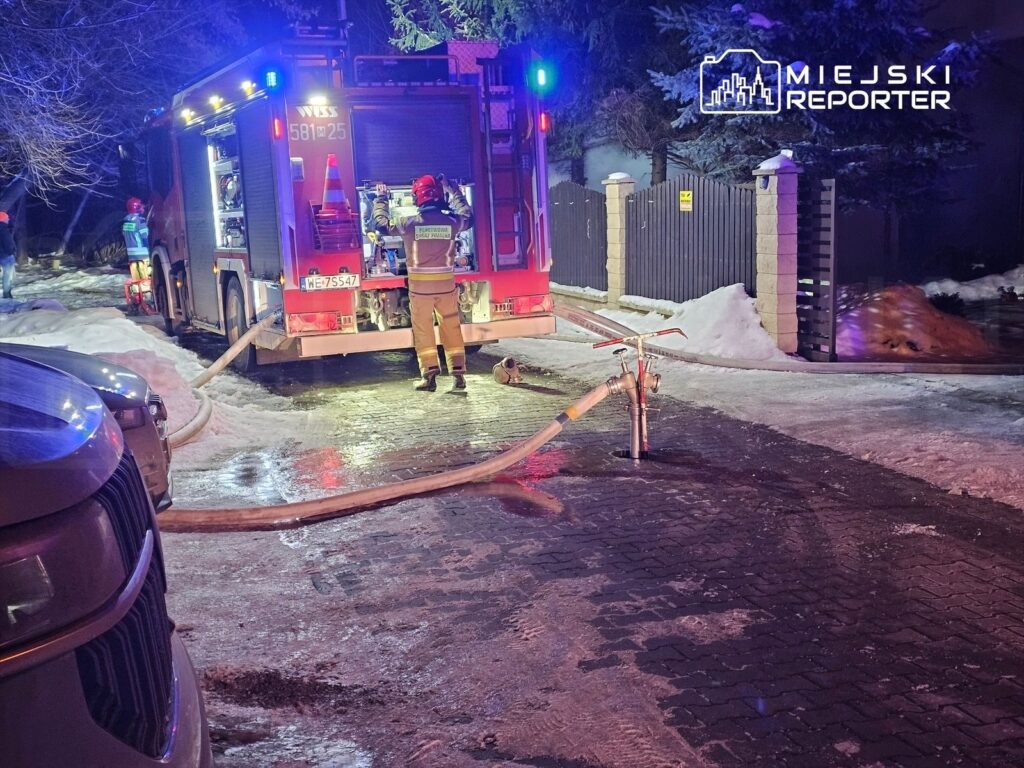 Strażacy przy wozie strażackim, podłączając wąż do hydrantu w oświetlonym nocą otoczeniu zaśnieżonej ulicy.