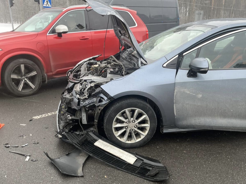 Zniszczony samochód osobowy z otwartą maską stoi na drodze, obok niego widoczny czerwony SUV. Na asfalcie leżą fragmenty karoserii.