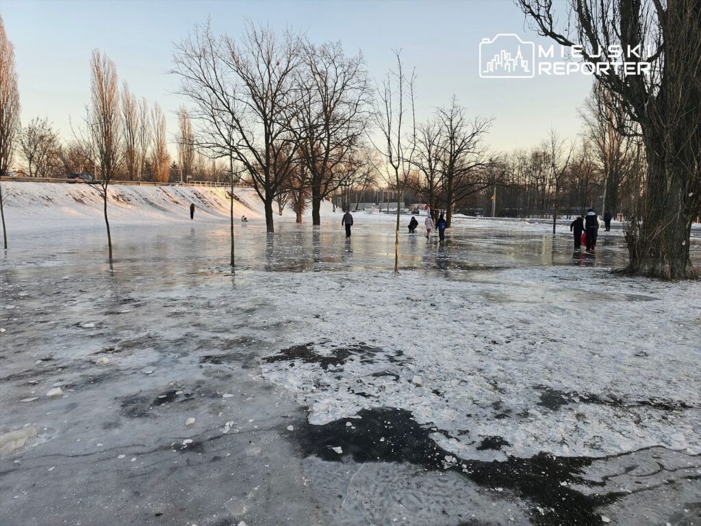 Grupa osób spaceruje po zamarzniętej powierzchni w parku, otoczonej nagimi drzewami i śniegiem.