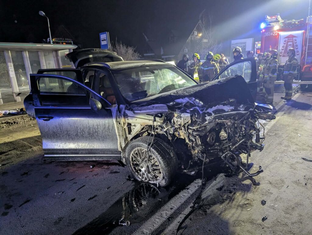 Uszkodzony samochód osobowy z rozbitą przednią częścią na drodze, obok grupy strażaków w akcji ratunkowej.