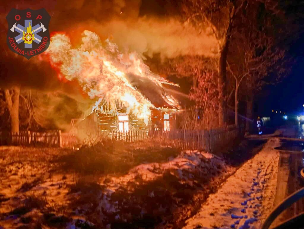 Płonący drewniany dom z ogniem wydobywającym się z dachu, otoczony przez drzewa i śnieg.