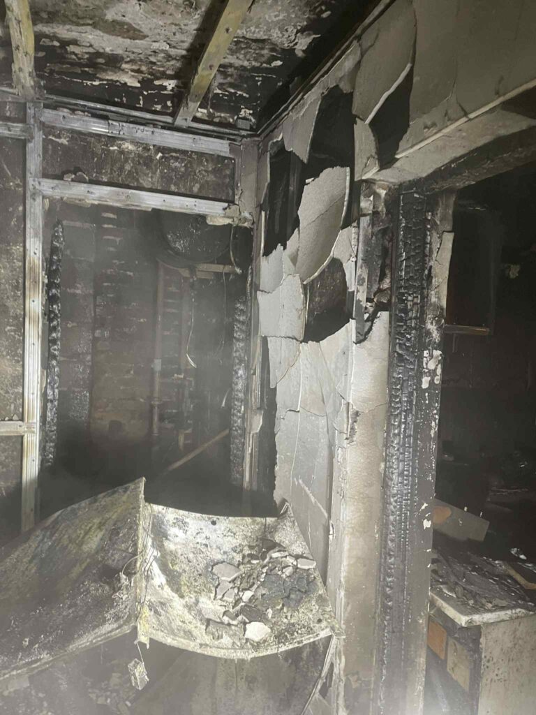 Zniszczona ściana w pomieszczeniu z odpadami po pożarze, z widocznymi spaloną farbą i uszkodzeniami strukturalnymi.